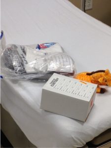 Chain of custody box for storage of evidence, clothes in Hefty zipper bag, and a child's toy on a hospital exam table